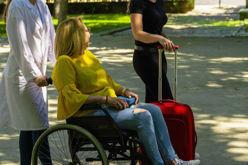 Woman in a wheelchair being pushed while talking with another woman with a suitcase on an out of focus background.