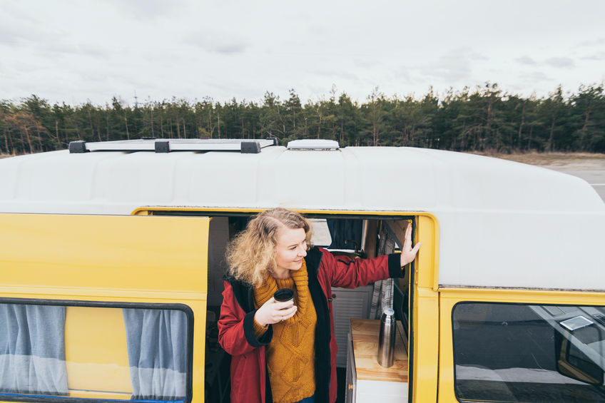 Young blond woman travelling by camper van with solar panel on the roof top and pine forest on the background